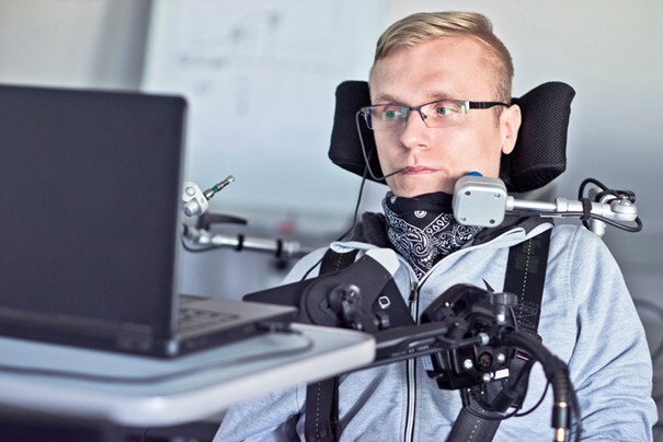 Mann mit schwerer körperlicher Behinderung nutzt diverse technische Hilfsmittel, um am PC arbeiten zu können.